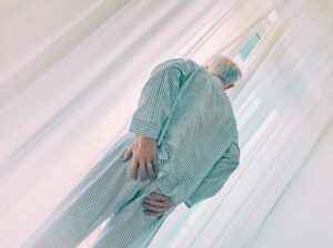 Older man in striped pajamas standing with a hand on his lower back beside sheer white curtains