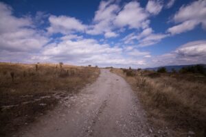Dusty gravel road winds through an open plain toward the horizon under a blue sky with scattered clouds