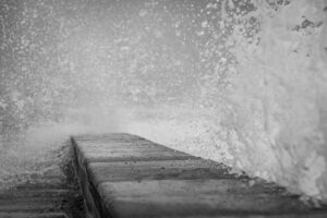 Nicolas houdayer uxibl6i7p9i unsplash | 日本白井新聞 Black and white photo of a concrete jetty with a large wave crashing against the edge spraying water into the air in front of it