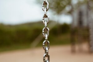 Close-up of a metal swing chain hanging from a playground, with a blurred playground backdrop.